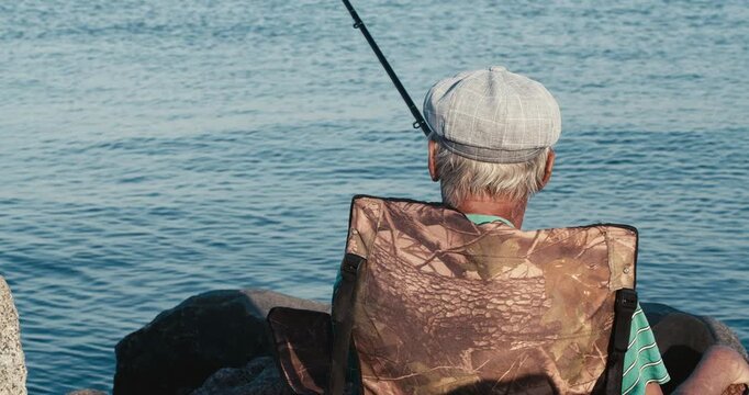 Close cinematic rear view of an elderly fisherman in a checked flat cap and vest holding his rod while fishing on a calm lake.