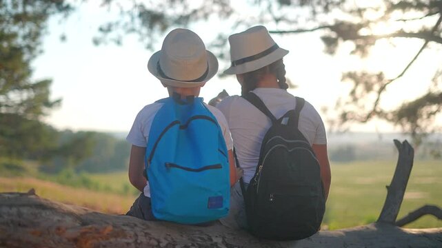 Boy and girl sitting on tree in nature. Summer outdoor relax with backpack. Children sit side by side wearing hat. Nature moment with children backpack. Girl and boy summer scene with backpack