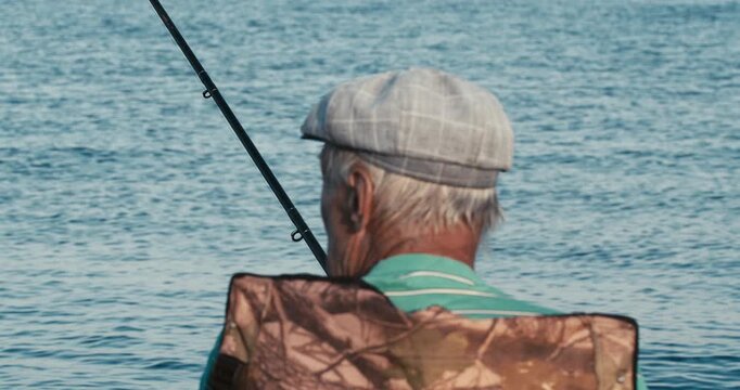 Cinematic rear view of an elderly fisherman in a flat cap sitting in a folding chair, holding a rod as he fishes on a calm lake.