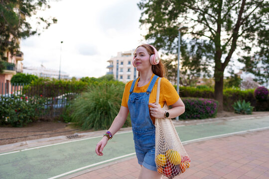 Woman walking outdoors listening to music and carrying fresh fruit