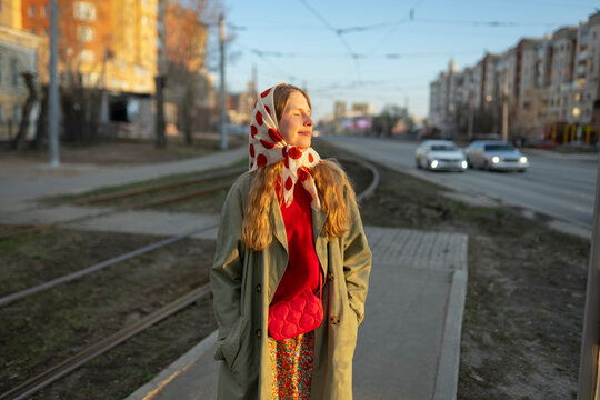 Pedestrian in raincoat and scarf enjoying sunlight at a bus stop