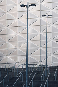 Modern architecture rises above stairs and railing beneath a geometric facade, where urban light poles emphasize pattern, scale and clean structure