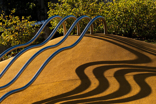 Curved blue railing crosses a yellow mound in an urban playground, where shadow and sunlight create abstract architecture beside dense greenery