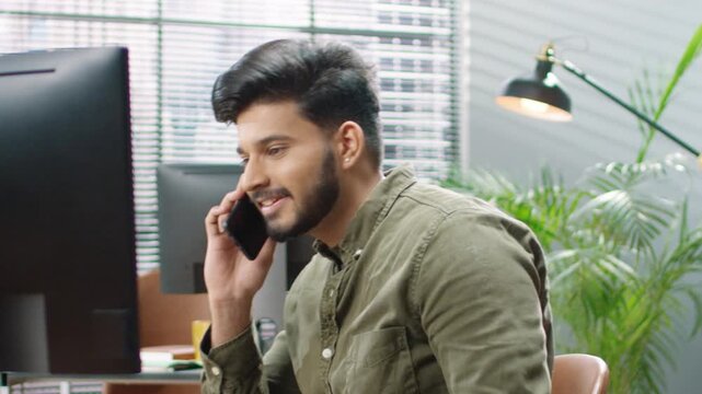 Smiling man in green shirt holding smartphone to ear, talking during phone call. Office with computer monitors, yellow mug, and indoor plant in background. Remotely speaking with someone.
