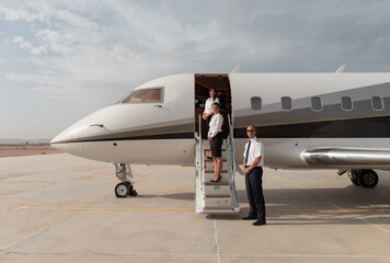 Confident private jet flight crew of two elegant flight attendants and a uniformed pilot standing on the airstairs of a luxury business jet on a sunlit desert airport tarmac at golden hour ideal for © .shock