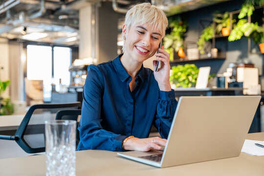 Smiling businesswoman talking on cell phone using laptop in office
