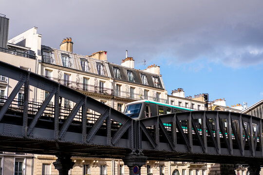 Paris metro moves across a steel bridge between classic buildings, where elevated rail transport cuts through an urban France street scene