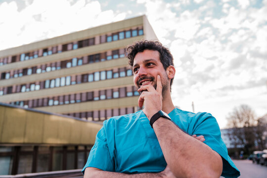 Smiling doctor in scrub thinking outside hospital building