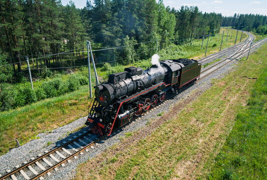 Aerial view captures an old black steam locomotive moving along parallel train tracks next to a dense green forest