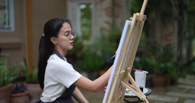 asian woman in casual white shirt holding brush painting artwork on canvas while enjoying relaxed moment in natural backyard garden space surrounded by green plants and creativity