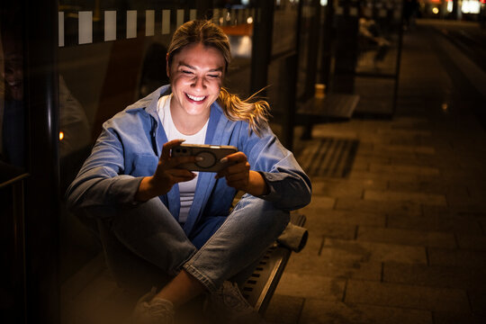 Woman smiling at smartphone waiting at city bus stop at night