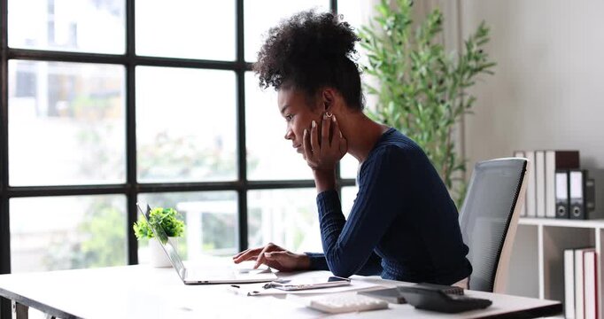 Focused young black businesswoman concentrating on laptop screen, working diligently on a project in a modern office setup, representing dedication, remote work, and career ambition