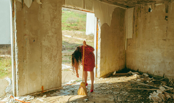 Exhausted person in red dress leans on broom in abandoned ruin
