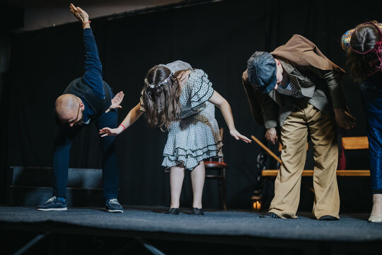 Young actors bow together at the front of a stage after finishing a play. The cast acknowledges the audience during a curtain call on a dark theater stage