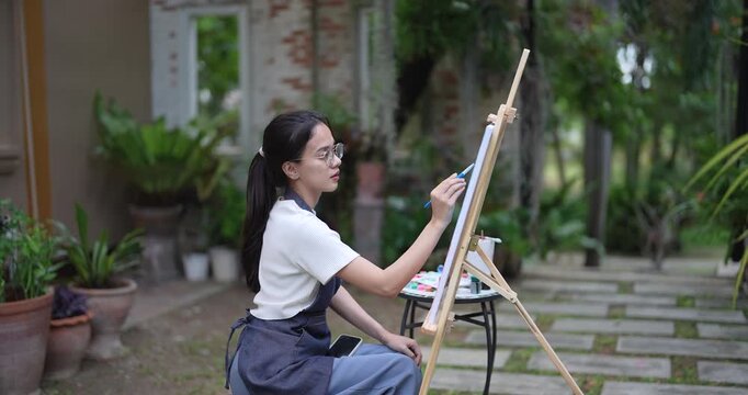 woman artist with long black hair sitting calmly while painting on canvas using brush expressing creative ideas in backyard filled with green potted plants and garden walkway
