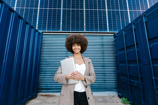 Smiling businesswomn holding laptop by solar panel and container