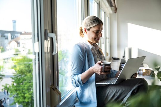 Smiling businesswoman working on laptop by the office window