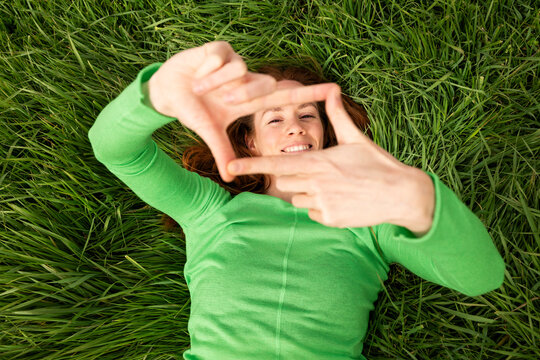 Smiling woman lying on grass framing face with hands outdoors
