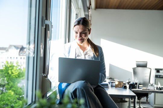 Smiling businesswoman working on notebook by window in modern office