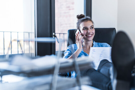 Smiling businesswoman talking on mobile phone with feet on desk