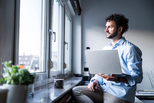 Smiling businessman holding laptop looking out office window