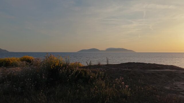 Aerial view of the rugged coastline with wildflowers and shrubs on a dirt path overlooking the sea and distant mountains at sunset in Zvernec, Vlore County, Albania.