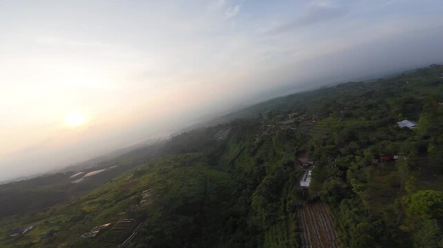 Aerial view of the sun rising over the lush green hills and tropical landscape near Joglo Java Neglasari in Cigombong, West Java, Indonesia.