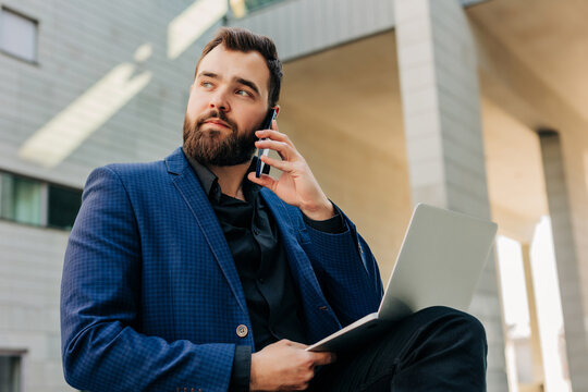 Executive in suit calling on smartphone outdoors with laptop