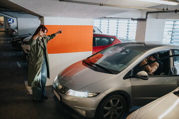 A woman gives a thumbs-up to a man seated in a silver car inside an indoor parking garage. She gestures to guide the driver as he maneuvers the vehicle into a parking space © qunica.com