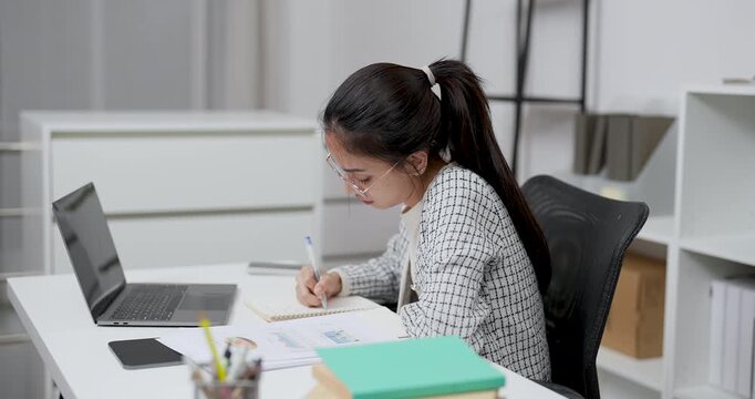 Asian young woman working alone in modern home office writing in notebook while looking at paperwork with laptop on desk focusing on planning work tasks in quiet organized workspace