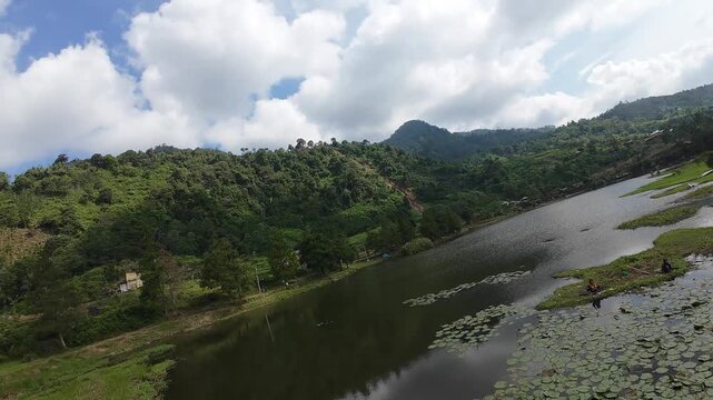 Aerial view of Rawa Gede Jonggol Lake with lily pads and lush green hills under a cloudy sky in Kecamatan Sukamakmur, Jawa Barat, Indonesia.