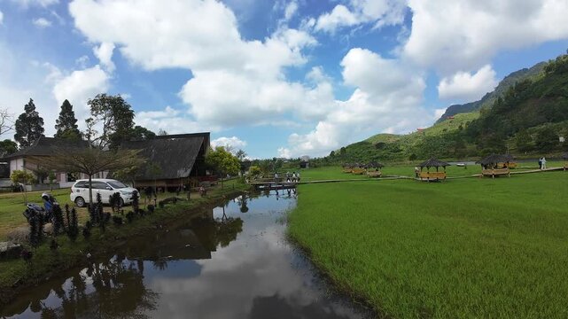 Aerial view of Rawa Gede Jonggol Lake with green fields, wooden gazebos, and mountains under a cloudy sky in Kecamatan Sukamakmur, Jawa Barat, Indonesia.