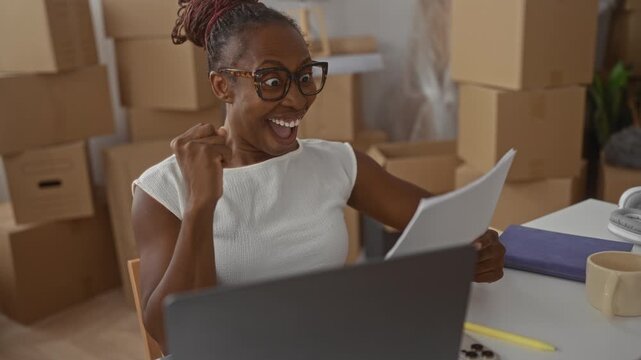 Woman holding paperwork and laptop, fist pump in an apartment filled with moving boxes; moving day joy.