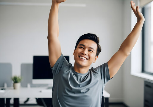 Smiling young Asian man in a modern office, happily stretching his arms above his head, enjoying a refreshing break from his professional workday