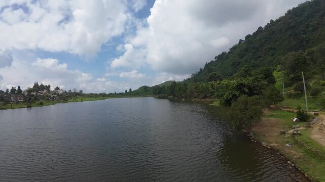 Aerial view of Rawa Gede Jonggol Lake surrounded by lush green hills and a cloudy sky in Kecamatan Sukamakmur, Jawa Barat, Indonesia.