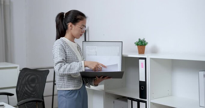 Asian businesswoman browsing through pages in large folder while standing beside cabinet in tidy home office environment reviewing important business documents with focus and care