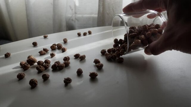 Hand pours dried tiger nuts from glass onto sunlit table, casual home setting