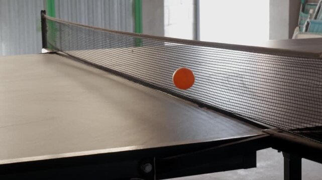 Orange table tennis ball bounces across net on empty indoor ping pong table