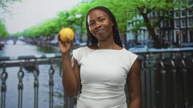 Young african american woman holds lemon with raised right hand on canal street bridge, slight head tilt and smirk visible; bemused wellness.