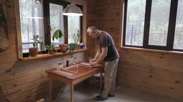 Man arranging wine glasses on dining table in cozy wooden cabin, preparing interior setting for meal service in rustic home environment