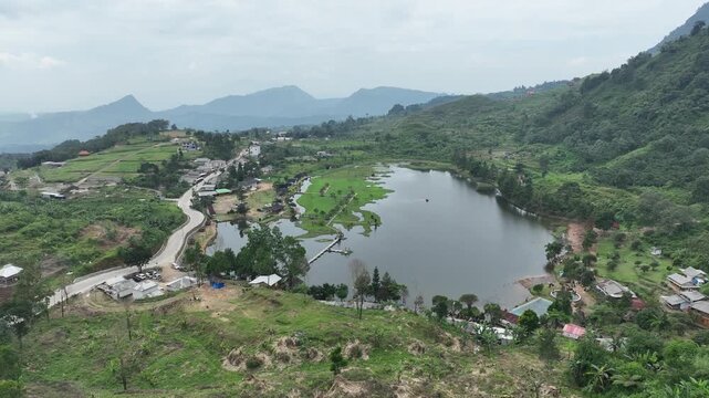 Aerial view of Rawa Gede Jonggol Lake surrounded by lush green hills, a small village, and a winding road in Kecamatan Sukamakmur, Jawa Barat, Indonesia.