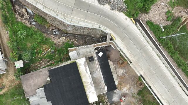 Aerial view of a curved concrete road winding past a building with a black roof and lush green vegetation near Rawa Gede Jonggol Lake Kecamatan Sukamakmur, Jawa Barat, Indonesia.