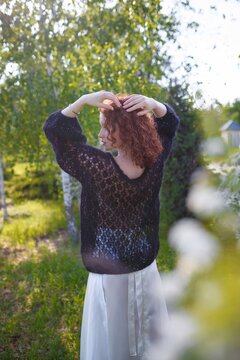 Redhead woman model in knitted sweater and silk skirt standing backwards on spring bloom background. Side view of face