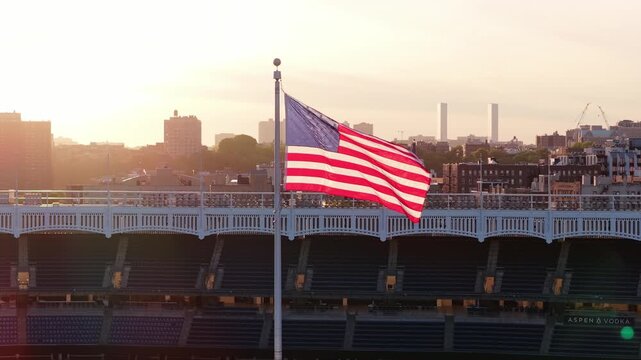American flag waving in wind with New York City South Bronx skyline, revealing empty Yankee Stadium seats and facade during warm and hazy sunset