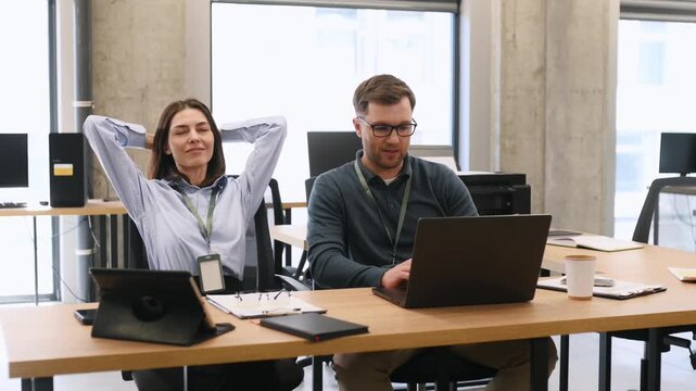 Caucasian woman in blue shirt stretches in office chair while male colleague works on laptop at shared workspace with modern furniture and bright lighting