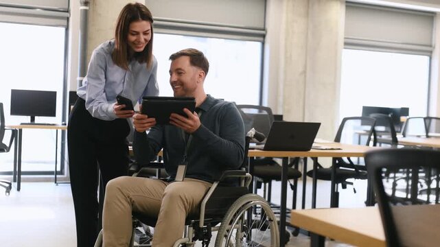 Businesswoman interacts with a man in a wheelchair using a laptop in a contemporary office environment featuring large windows and wooden furniture