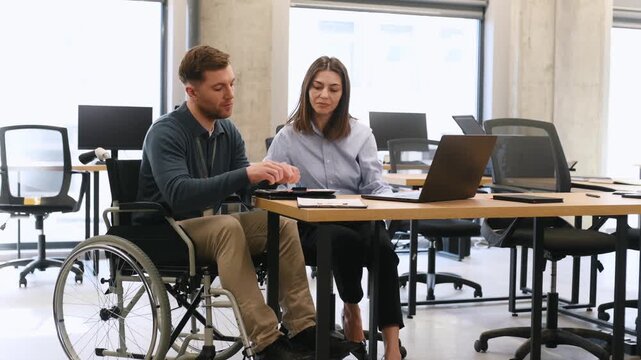 Young man in wheelchair and woman in professional attire discuss documents at a desk with a laptop in a contemporary office setting with empty desks