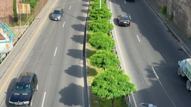 Cars and trucks travel along a multi-lane highway, with a central median of lush green trees separating the opposing directions of traffic under a bright sky. Armenia, Yerevan, 