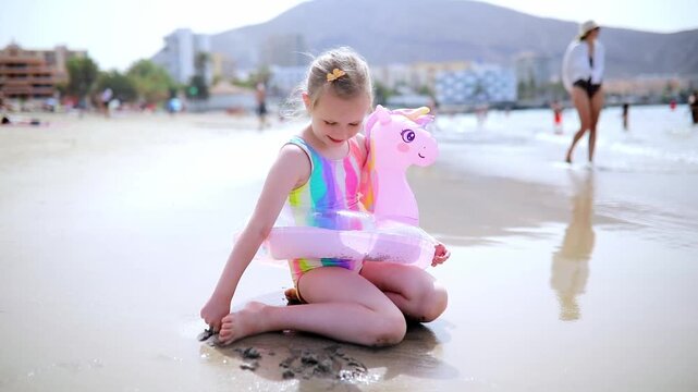 Little girl playing in shallow sea water with an inflatable unicorn swim ring in Los Cristianos, Tenerife, Canary Islands, Spain