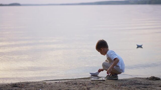 Crouching boy placing paper boat on calm lake water. Side view of child squatting at sandy shore launching origami boat at dusk.